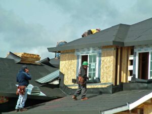 A homeowner reviews roofing estimates and loan documents at a dining table, with a shingle sample and mug nearby.