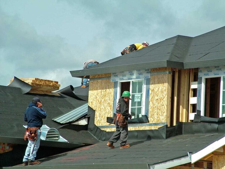 A homeowner reviews roofing estimates and loan documents at a dining table, with a shingle sample and mug nearby.